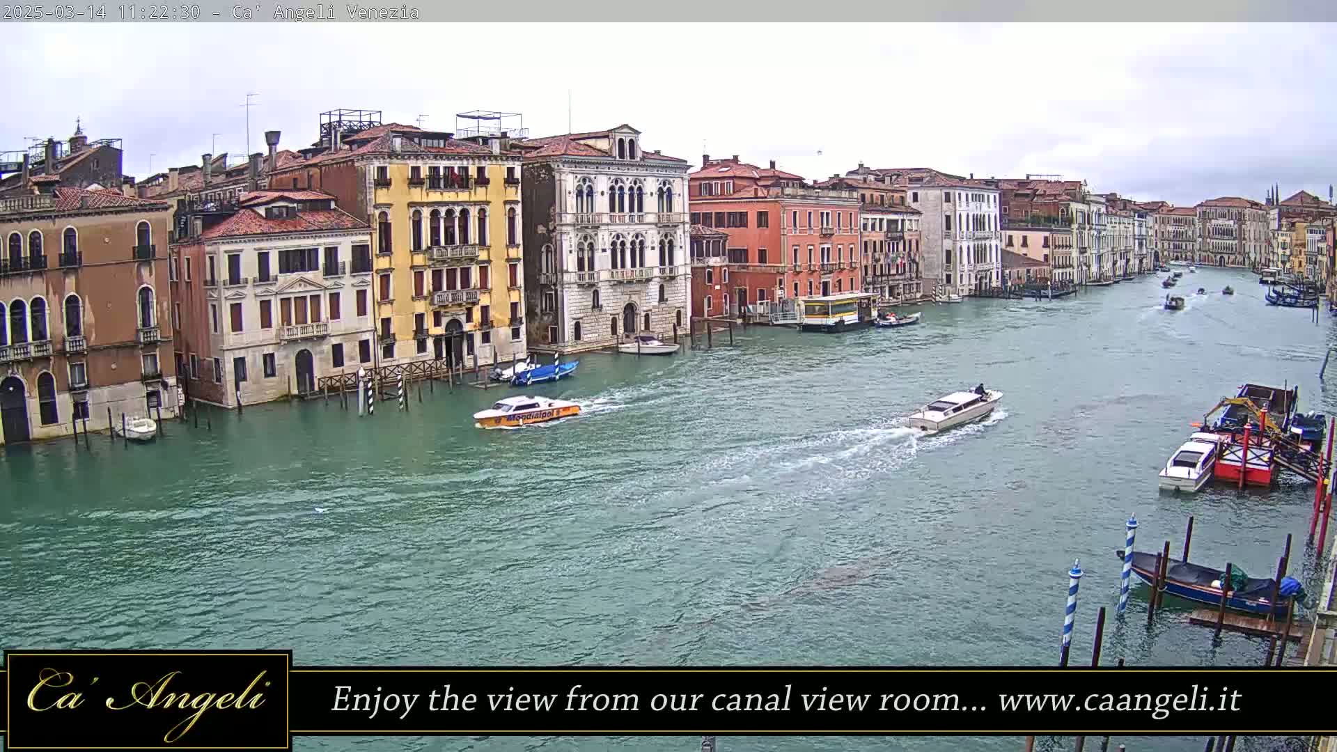 A wide shot shows a waterway in Venice, Italy on an overcast day with several boats traveling down the canal, and multiple buildings lining the waterway.