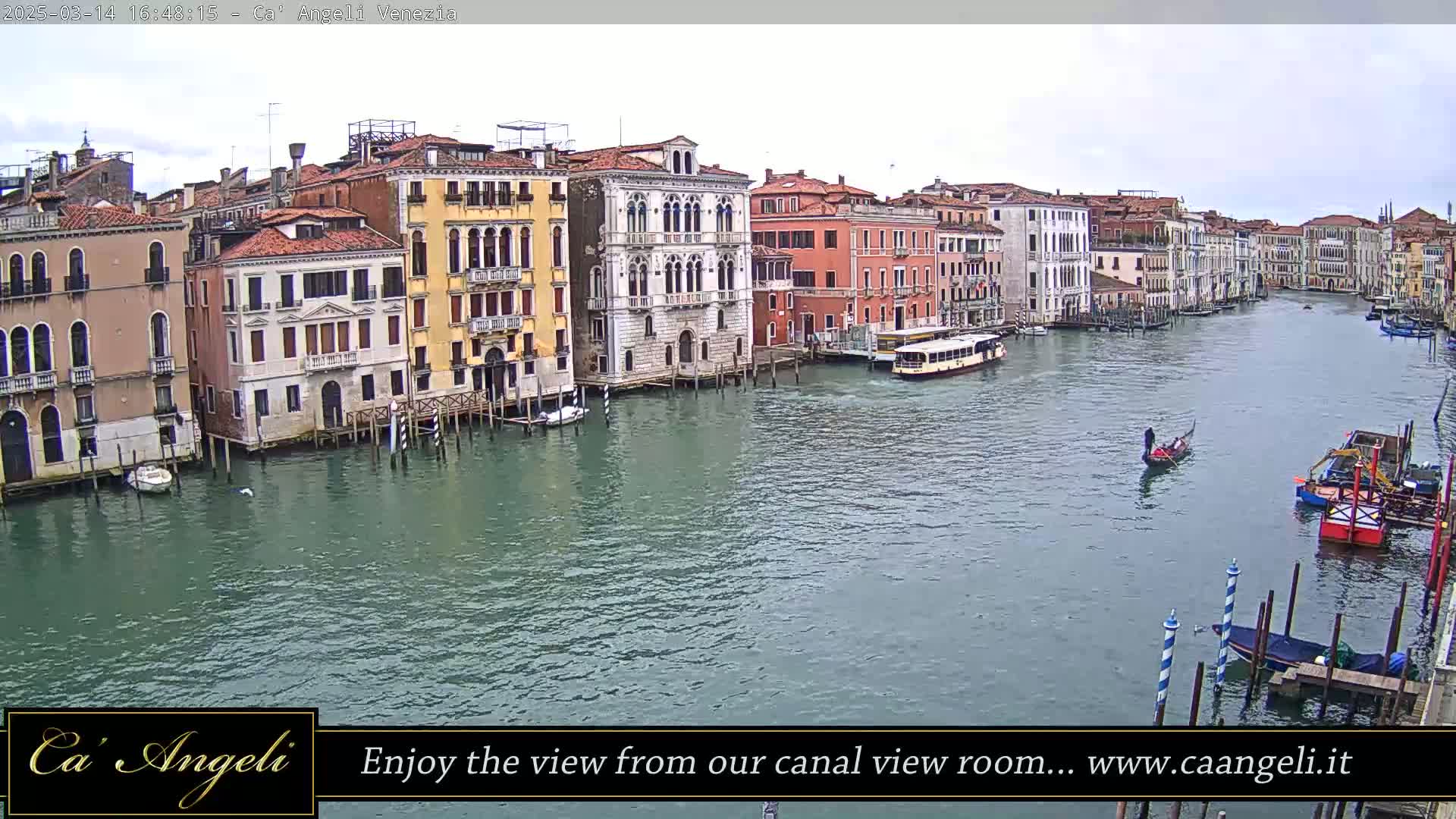 A wide shot of a canal in Venice, Italy, shows several buildings lining the waterway under an overcast sky, with boats including a gondola and a water bus on the water.