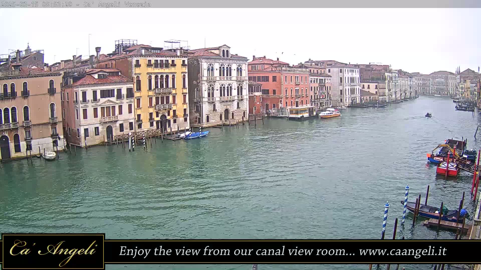 A wide shot of a canal in Venice on an overcast day shows numerous buildings lining the waterway with several boats moored and traveling along the canal.