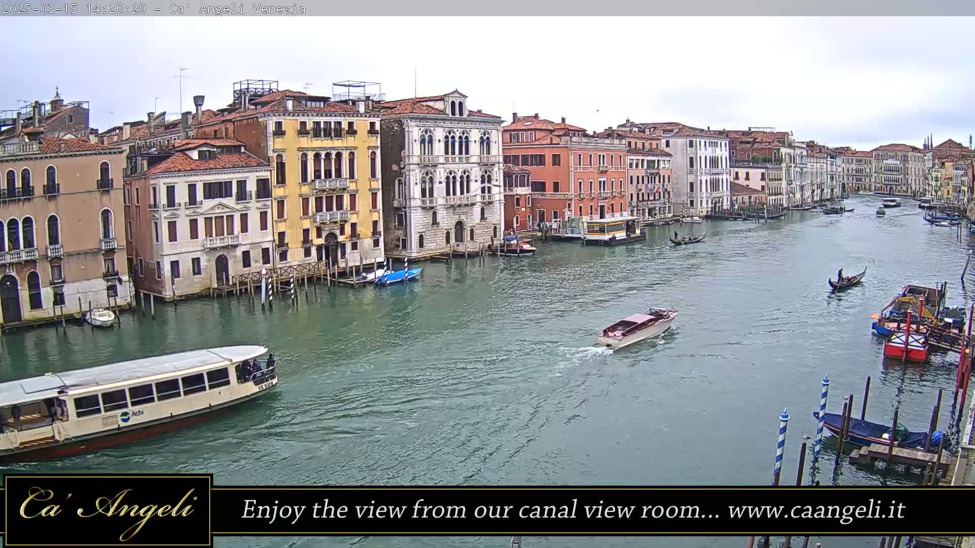 A waterway in Venice, Italy, shows several buildings along its edges and various boats, including gondolas and a water bus, under an overcast sky.