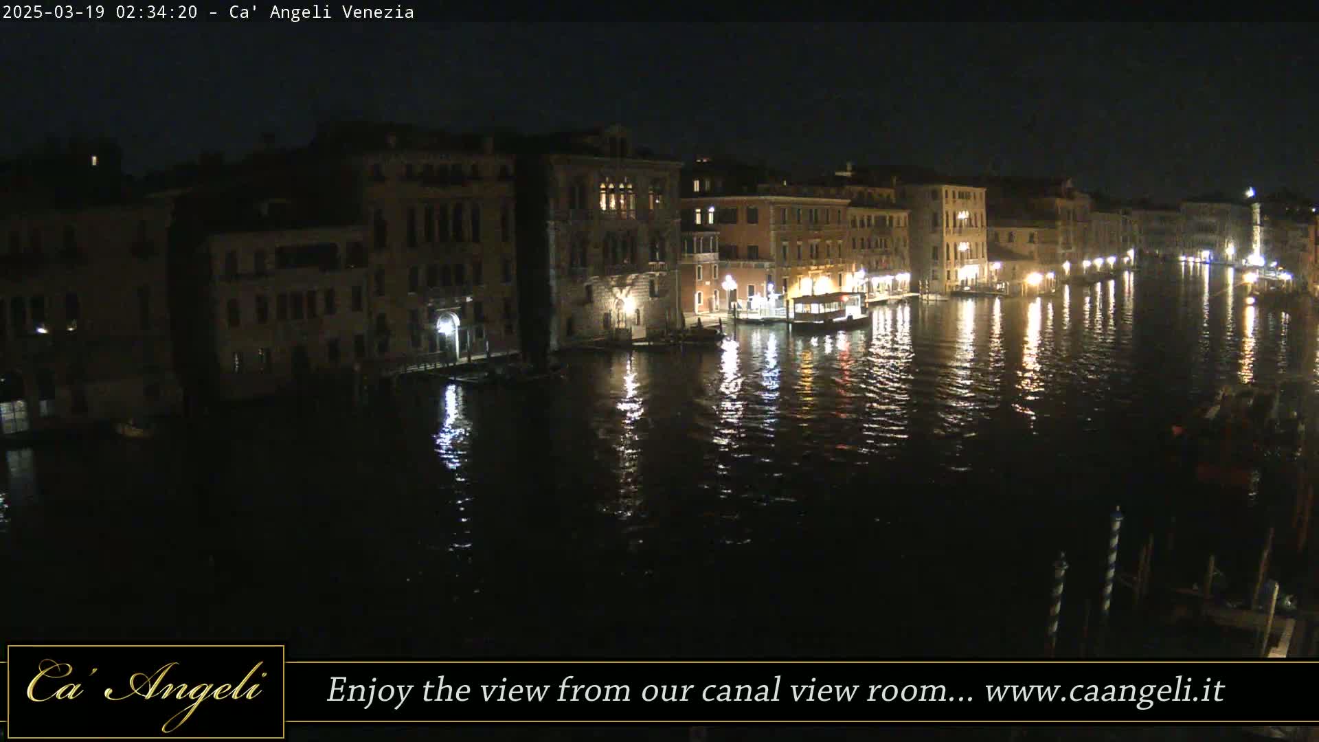 A nighttime view of a canal in Venice, Italy, shows illuminated buildings lining the waterway, with their lights reflecting on the calm water.