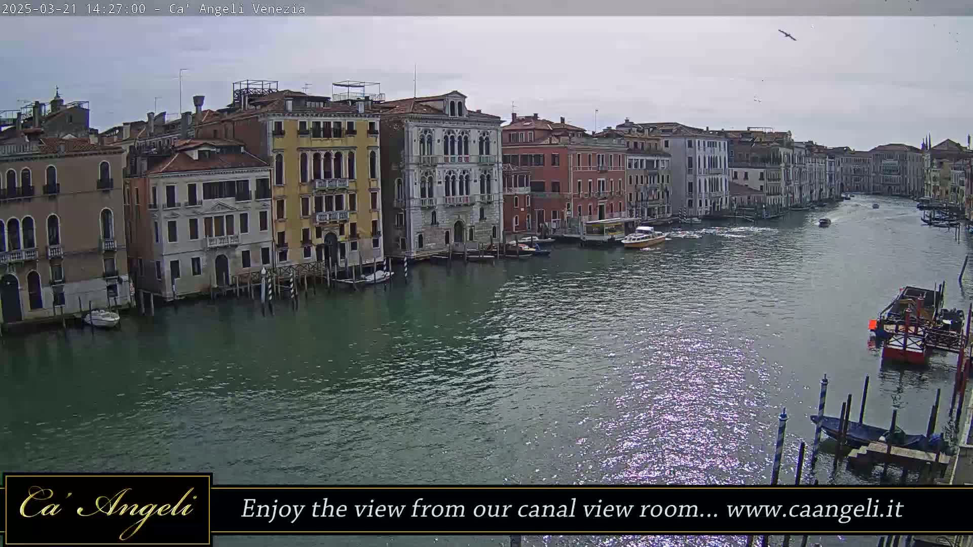 A wide shot shows a Venetian canal under a mostly cloudy sky, with several boats and colorful buildings lining the waterway.