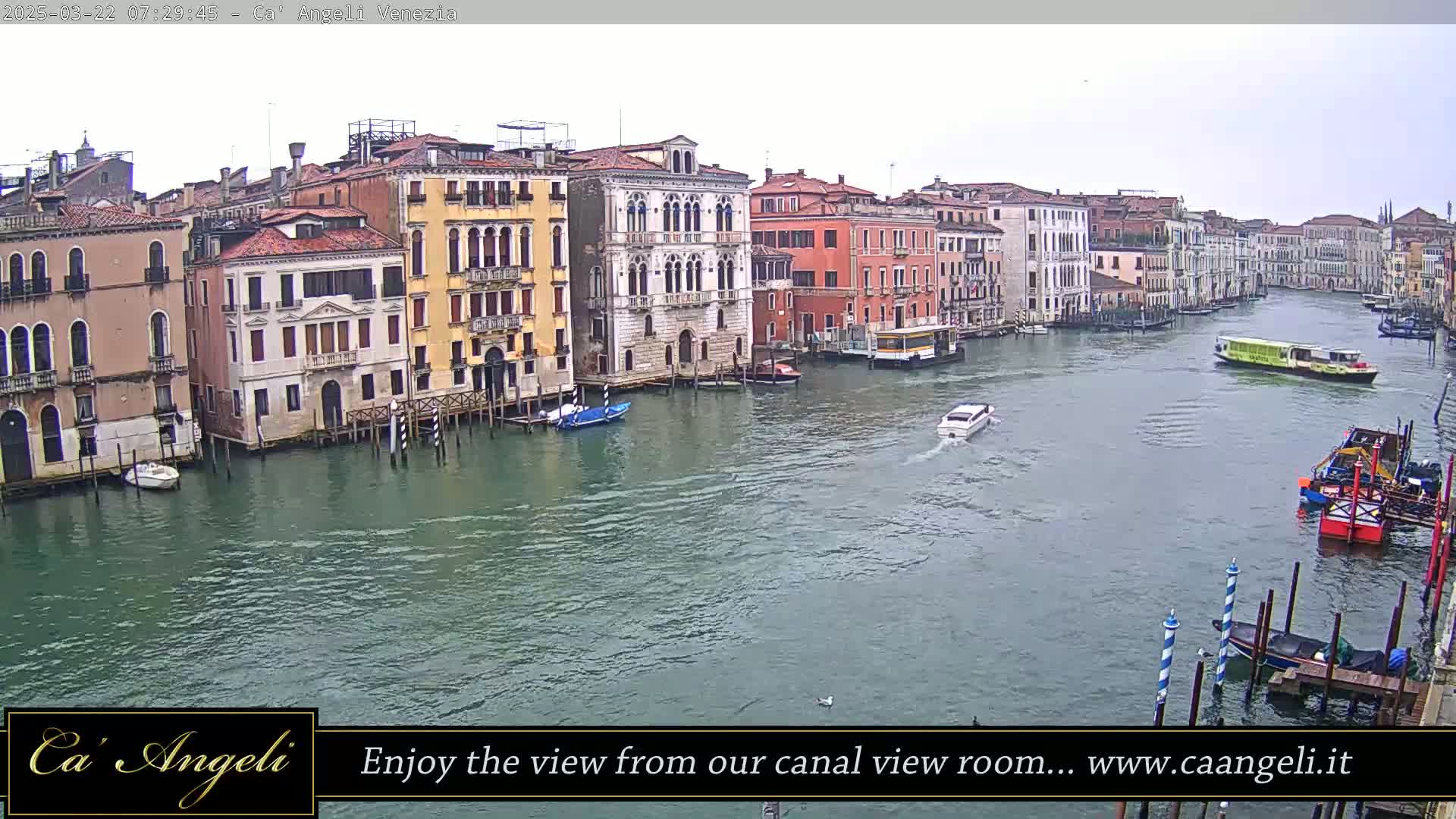 A wide shot shows a waterway in Venice with numerous buildings lining the banks on a cloudy day, several boats are visible on the water.