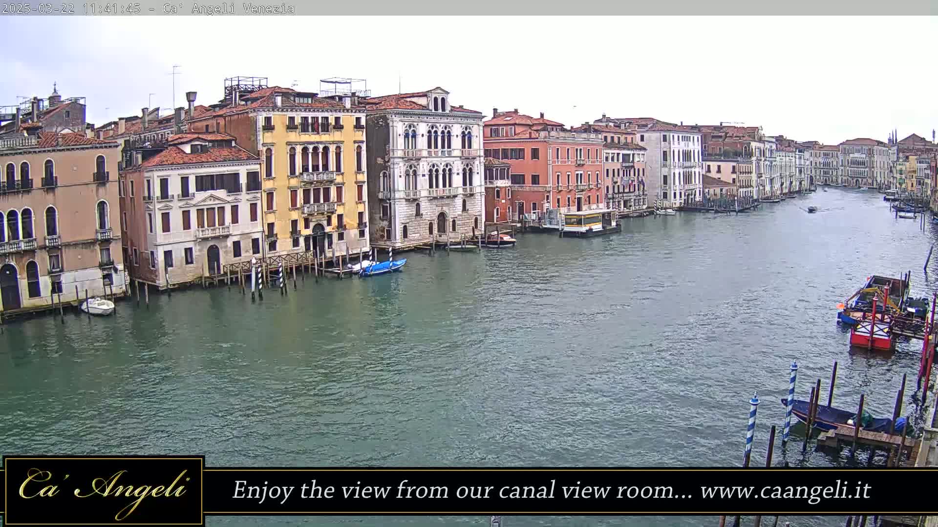 A wide shot of a canal in Venice, Italy, on an overcast day, shows several historic buildings lining the waterway with boats moored along the shore.