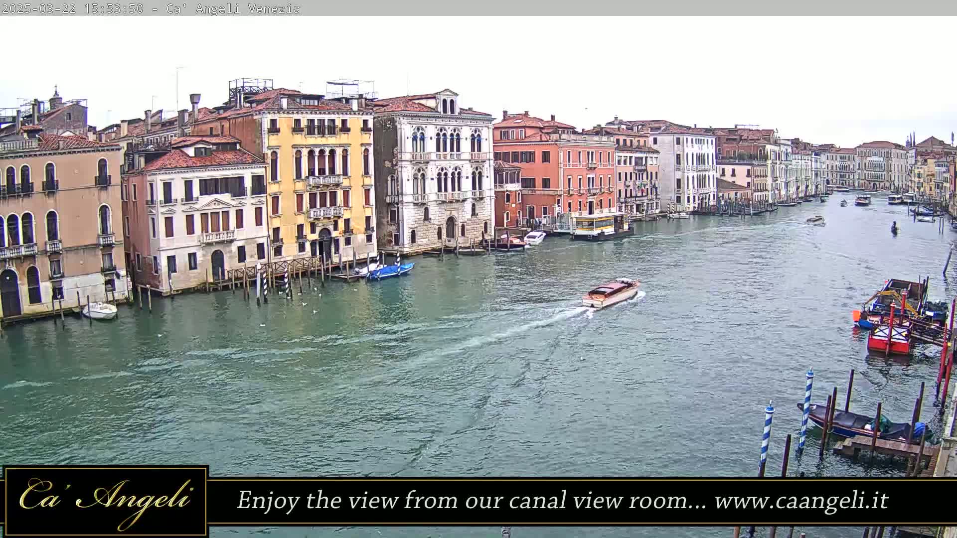 A wide shot of a waterway in Venice, Italy, on an overcast day, shows several boats and numerous buildings lining the canal.