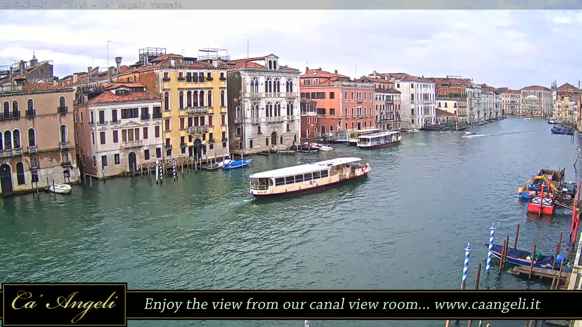 A wide shot shows a waterway with several boats and numerous buildings lining the banks under an overcast sky.