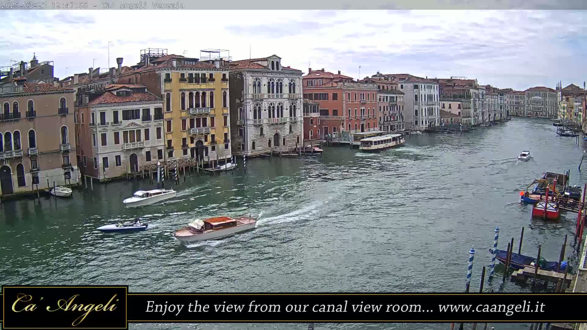 Several motorboats travel along a wide canal lined with closely-packed, multi-story buildings under a cloudy sky.