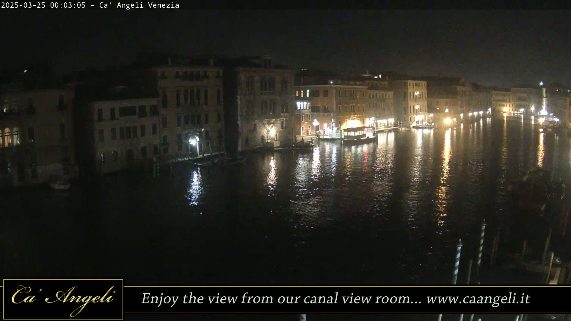 A nighttime view of a canal in Venice, Italy, shows illuminated buildings lining the waterway, with boats moored along the banks and their lights reflecting on the dark water.