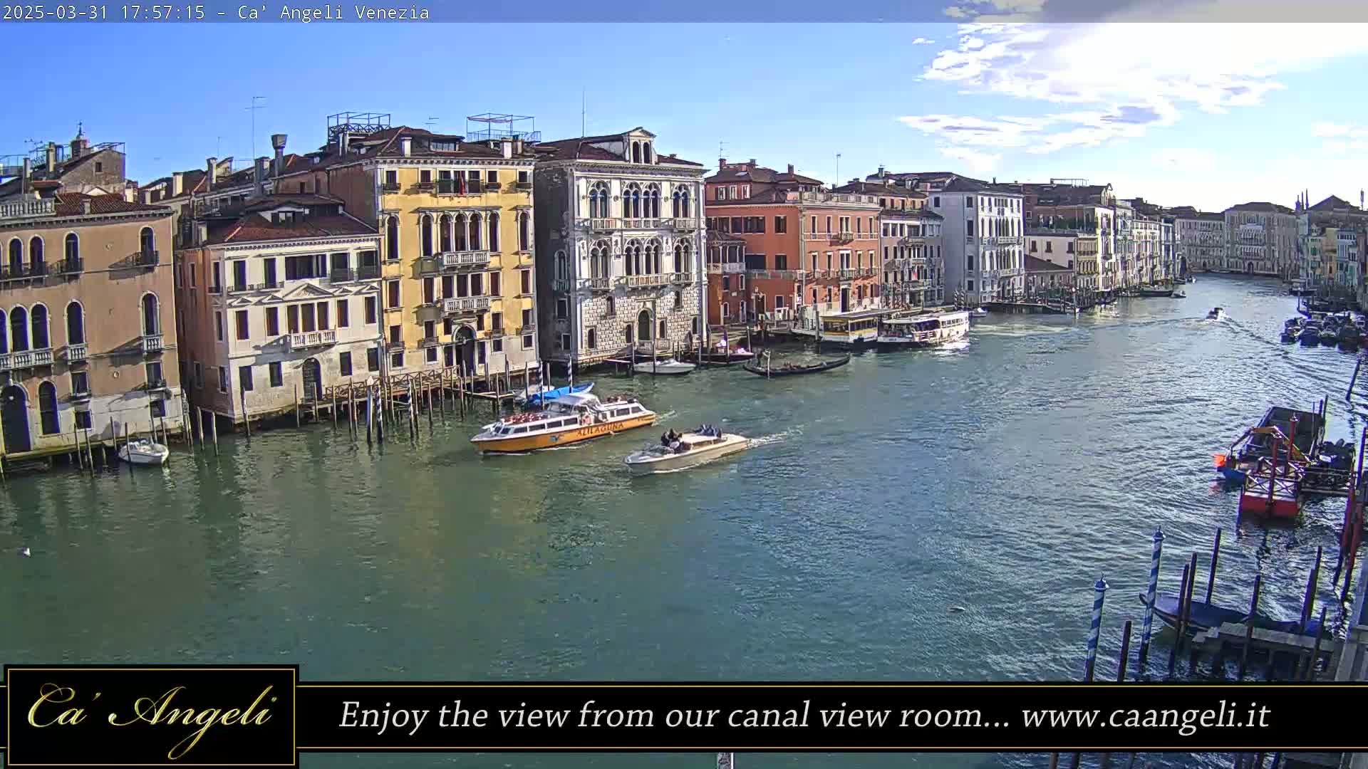 A sunny day in Venice shows a wide canal with several boats and numerous buildings lining the waterway.