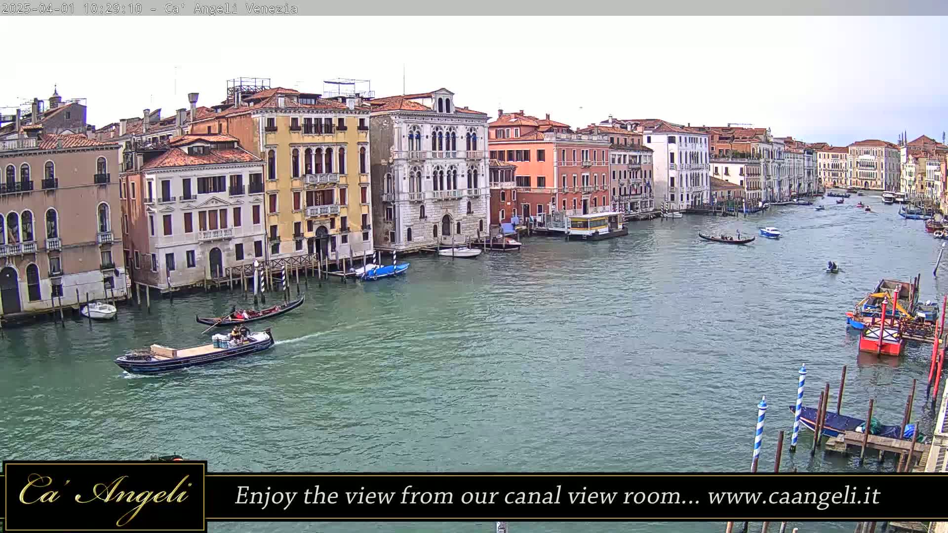 A canal in Venice, Italy, shows numerous buildings lining the waterway, with gondolas and other small boats traveling on the calm, green water under an overcast sky.