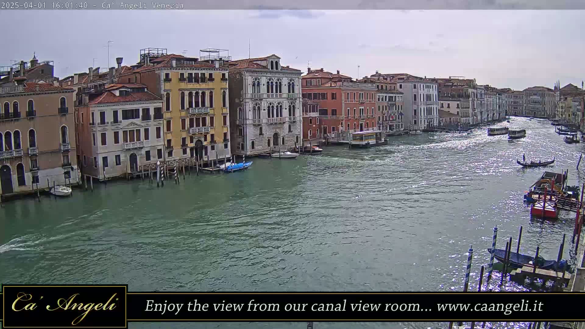 A canal in Venice, under a mostly cloudy sky, is filled with boats and lined with tall, colorful buildings.