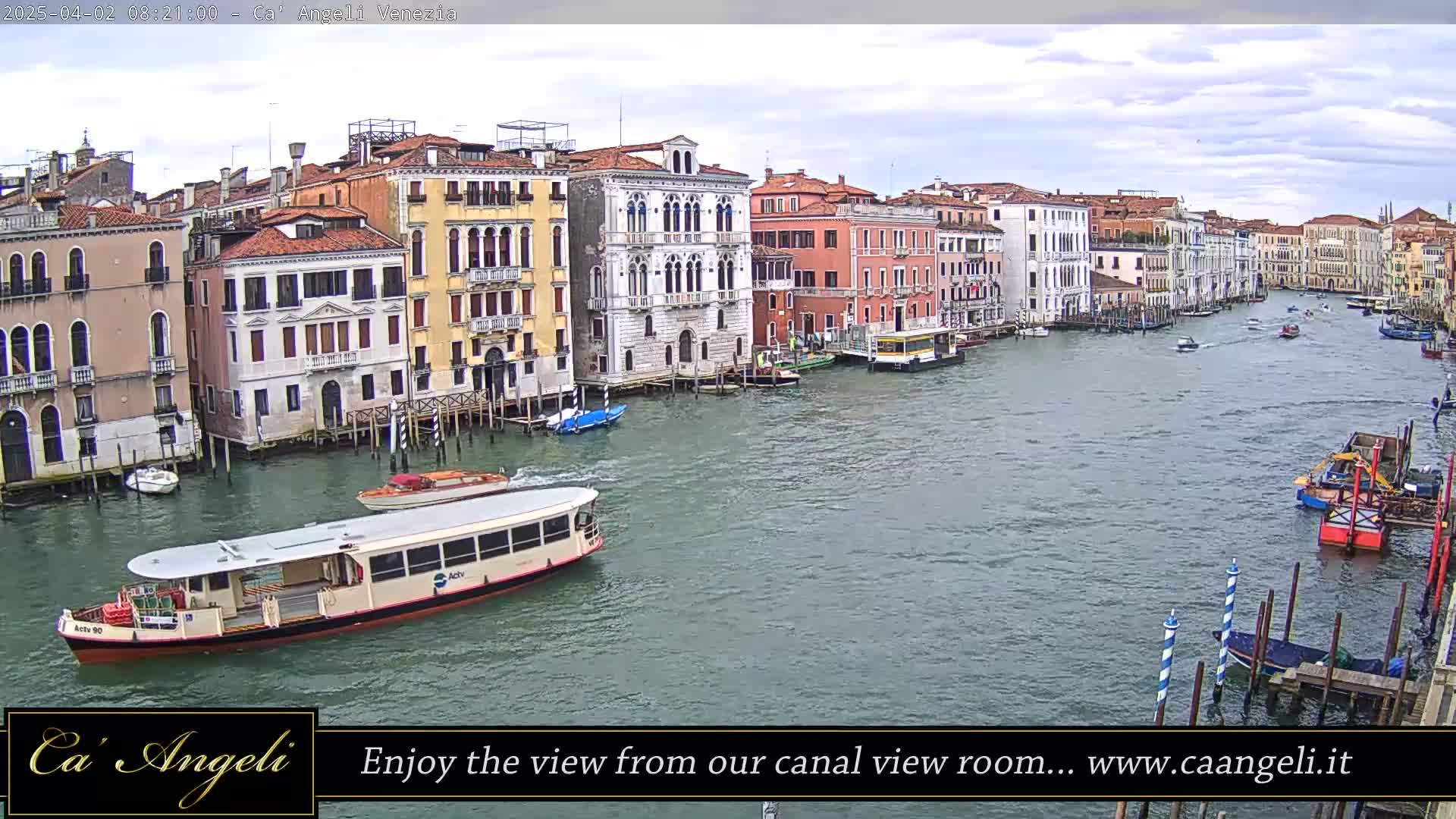 A wide canal in Venice, under a cloudy sky, is filled with boats, including a water bus, and lined with closely spaced buildings.