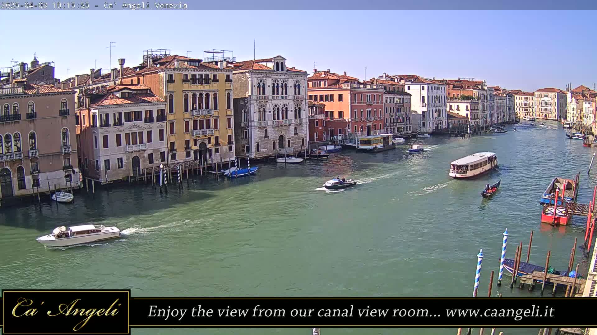 A sunny day in Venice shows a wide canal with several boats, gondolas, and water taxis passing by colorful buildings lining the waterway.