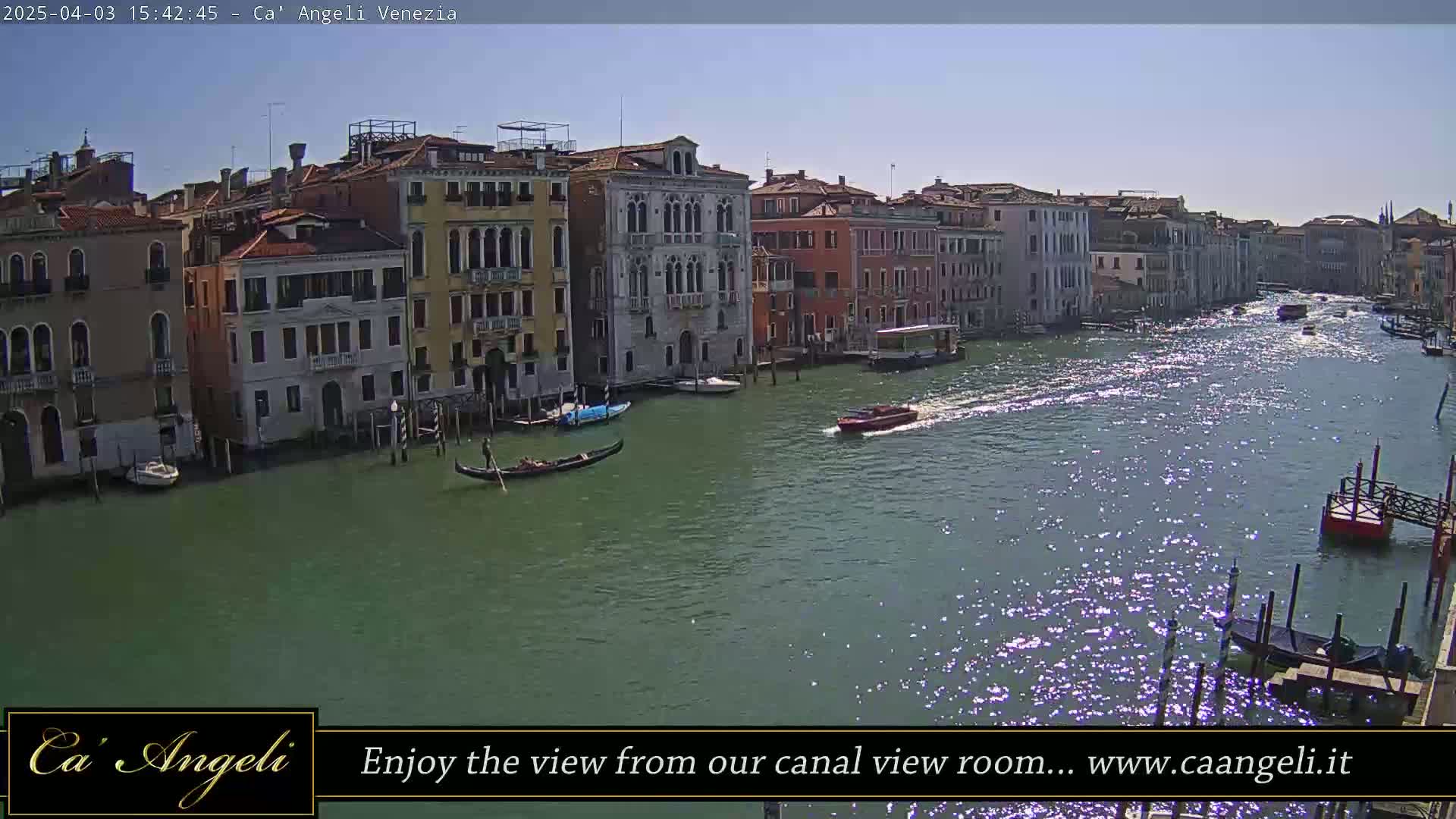 A sunny day in Venice shows a canal with gondolas and motorboats passing by colorful buildings lining the waterway.