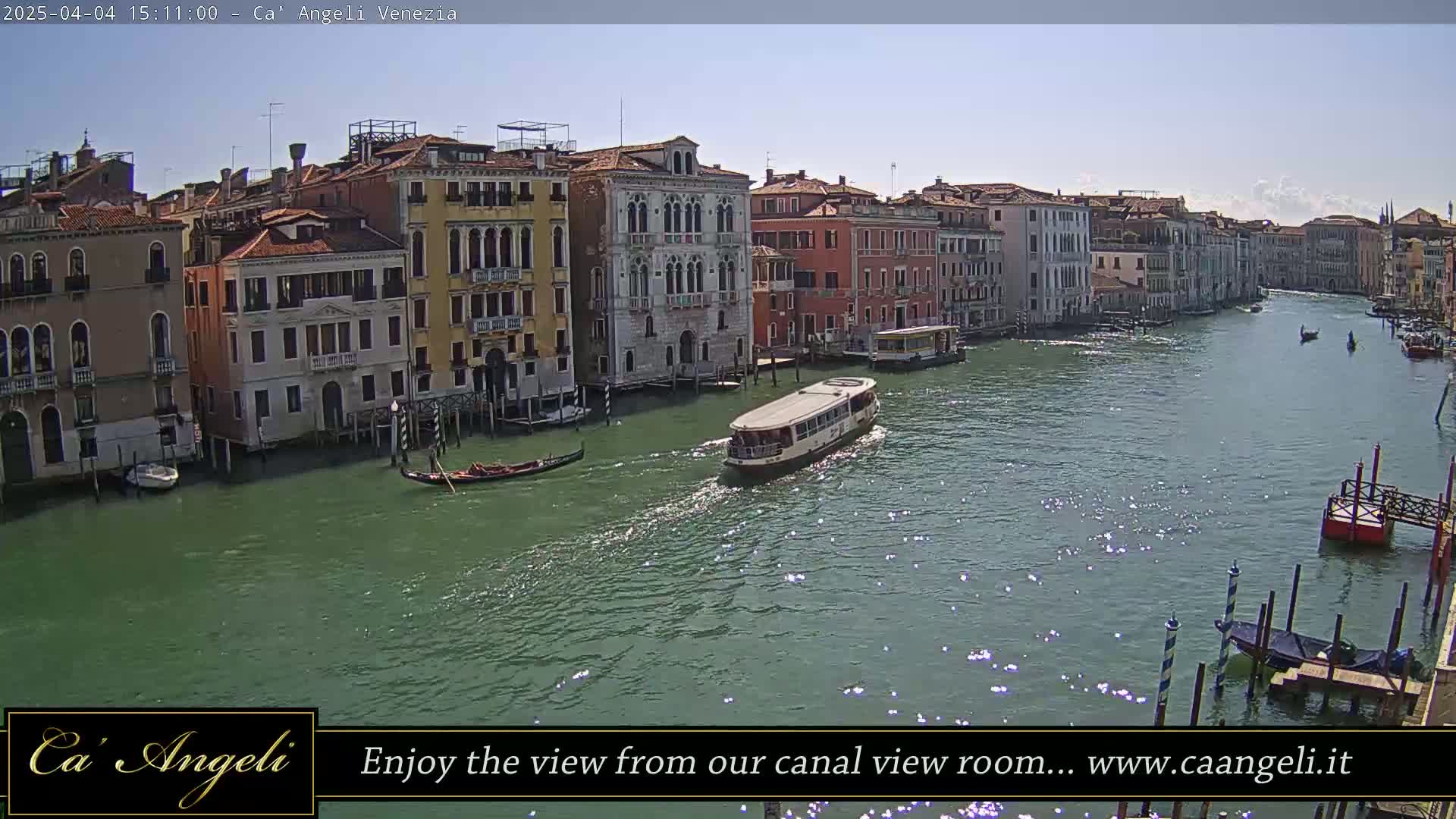 Under a sunny sky, a waterway flows past colorful buildings in Venice, with gondolas and a water bus visible on the water.