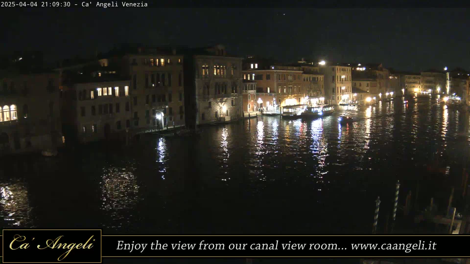 A nighttime view of a canal in Venice, Italy, shows several buildings with lights on along the waterway, and boats are moored along the banks.