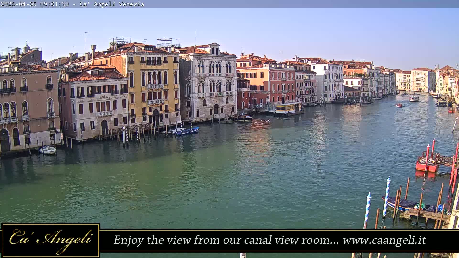 A wide shot of a canal in Venice, Italy, on a sunny day, shows numerous buildings lining the waterway with boats moored along the edges.