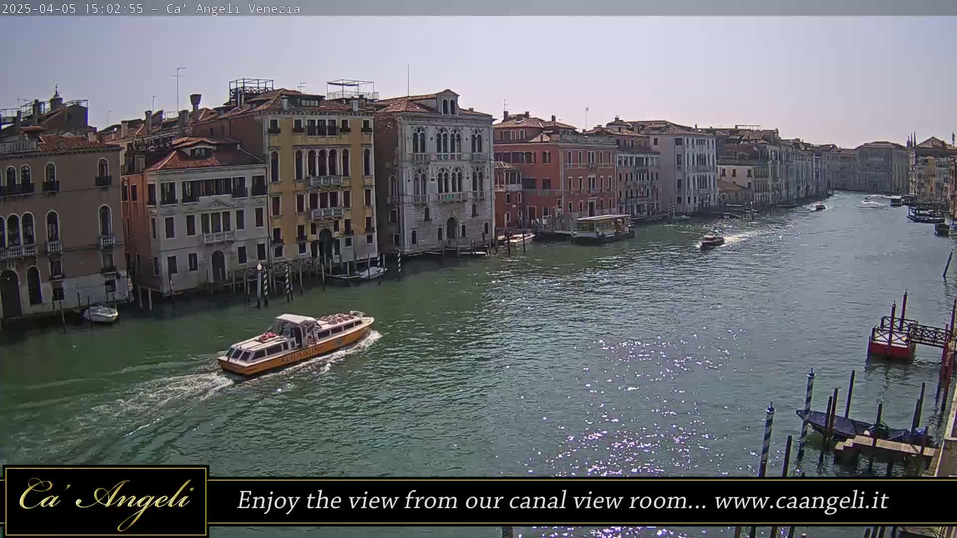 A sunny day in Venice shows a waterway with several buildings lining the canal, and a yellow water taxi travels down the center.
