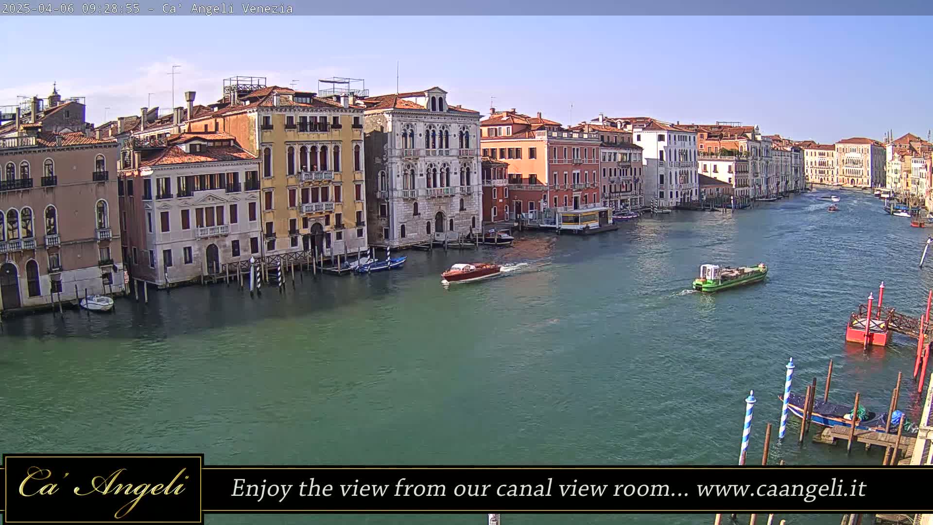 A sunny day in Venice shows a wide canal with colorful buildings lining the banks, several small boats traveling on the water, and a few docked near the bottom of the frame.