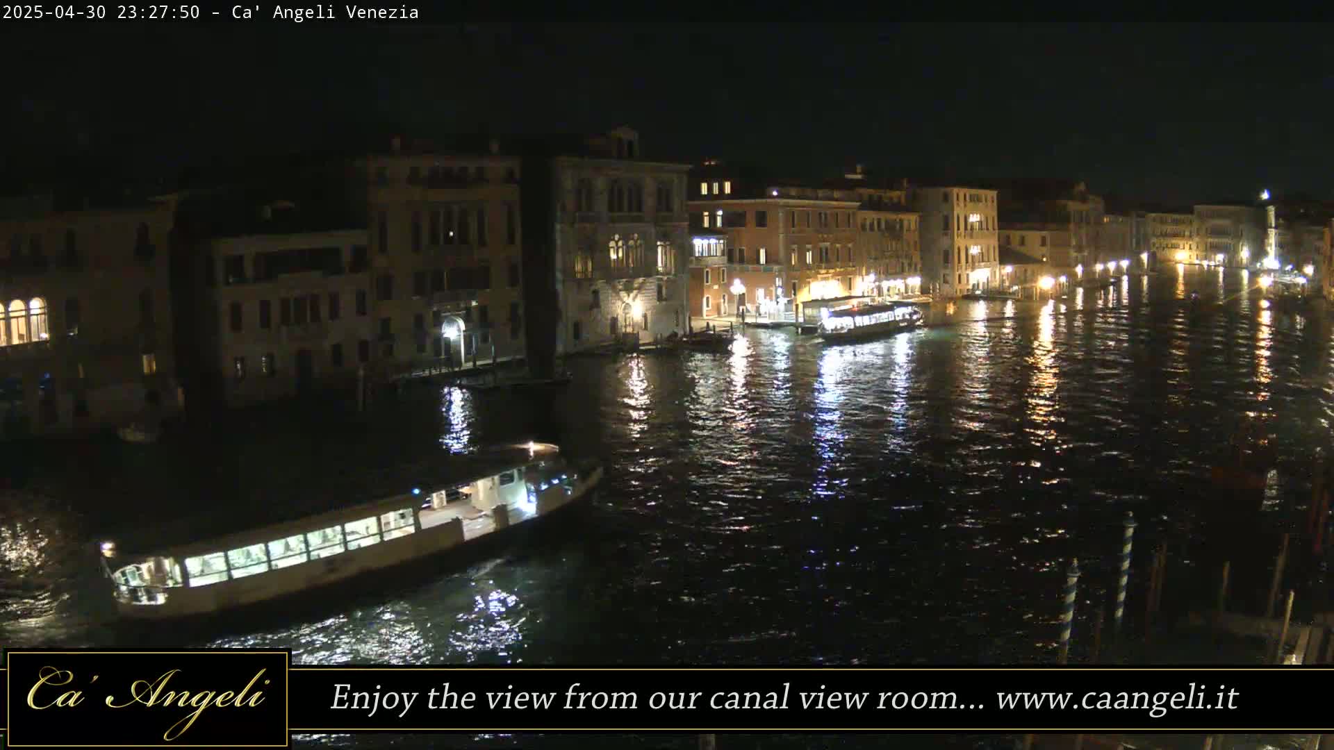 A nighttime view of a canal in Venice, Italy, shows illuminated buildings lining the waterway and several boats on the water.