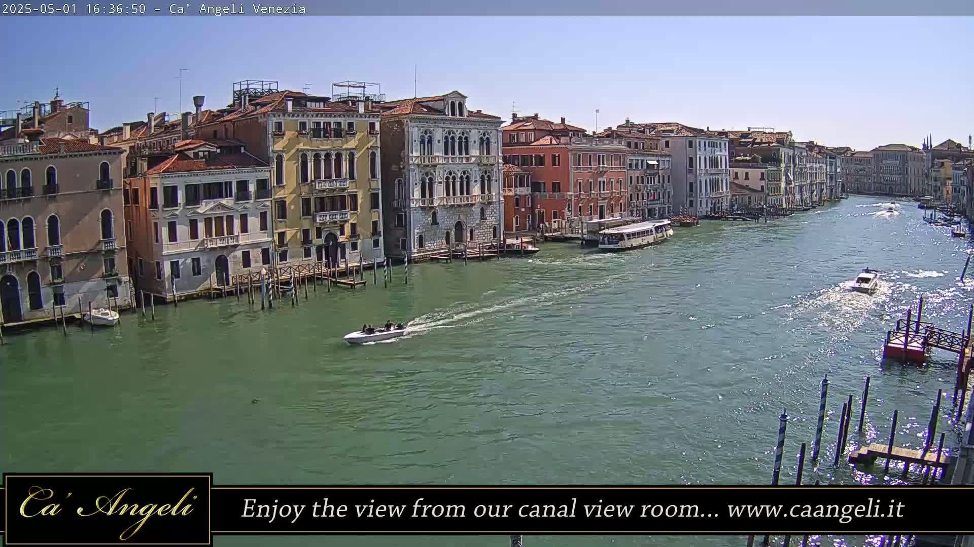 A sunny day in Venice shows a wide canal with several boats passing by colorful buildings lining the waterway.