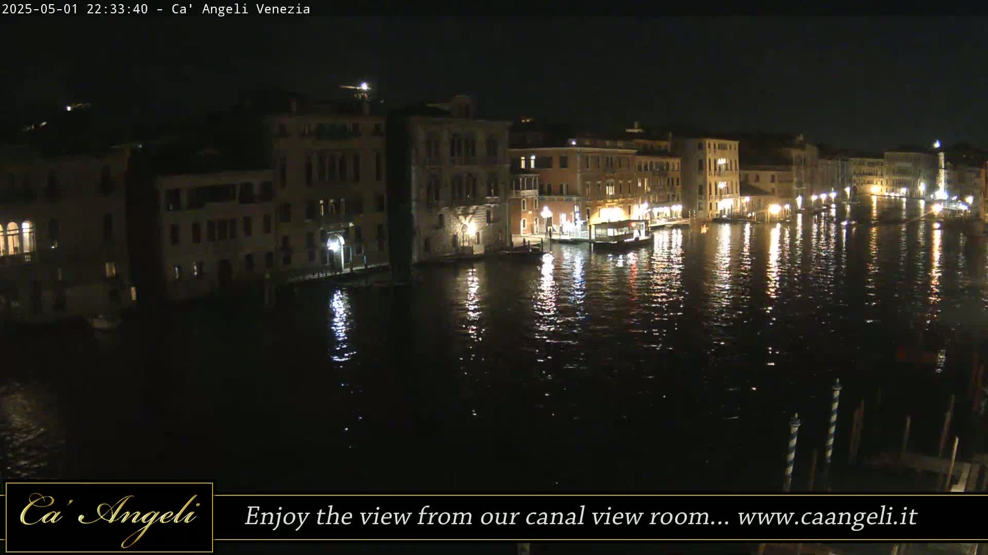 A nighttime view of a canal in Venice, Italy, shows illuminated buildings lining the waterway, their lights reflecting on the dark water.