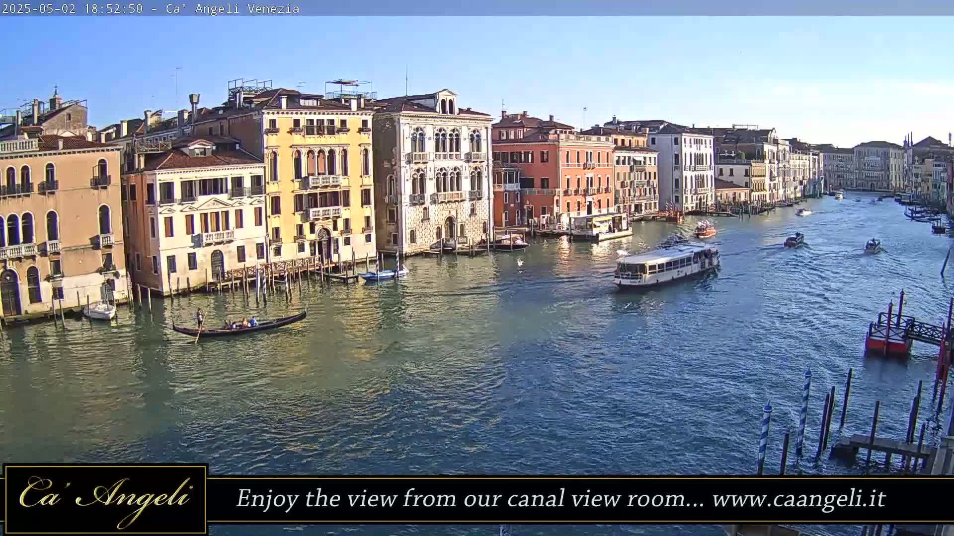 A sunny day in Venice shows a wide canal with several boats, including a gondola and a water bus, passing colorful buildings along the waterfront.