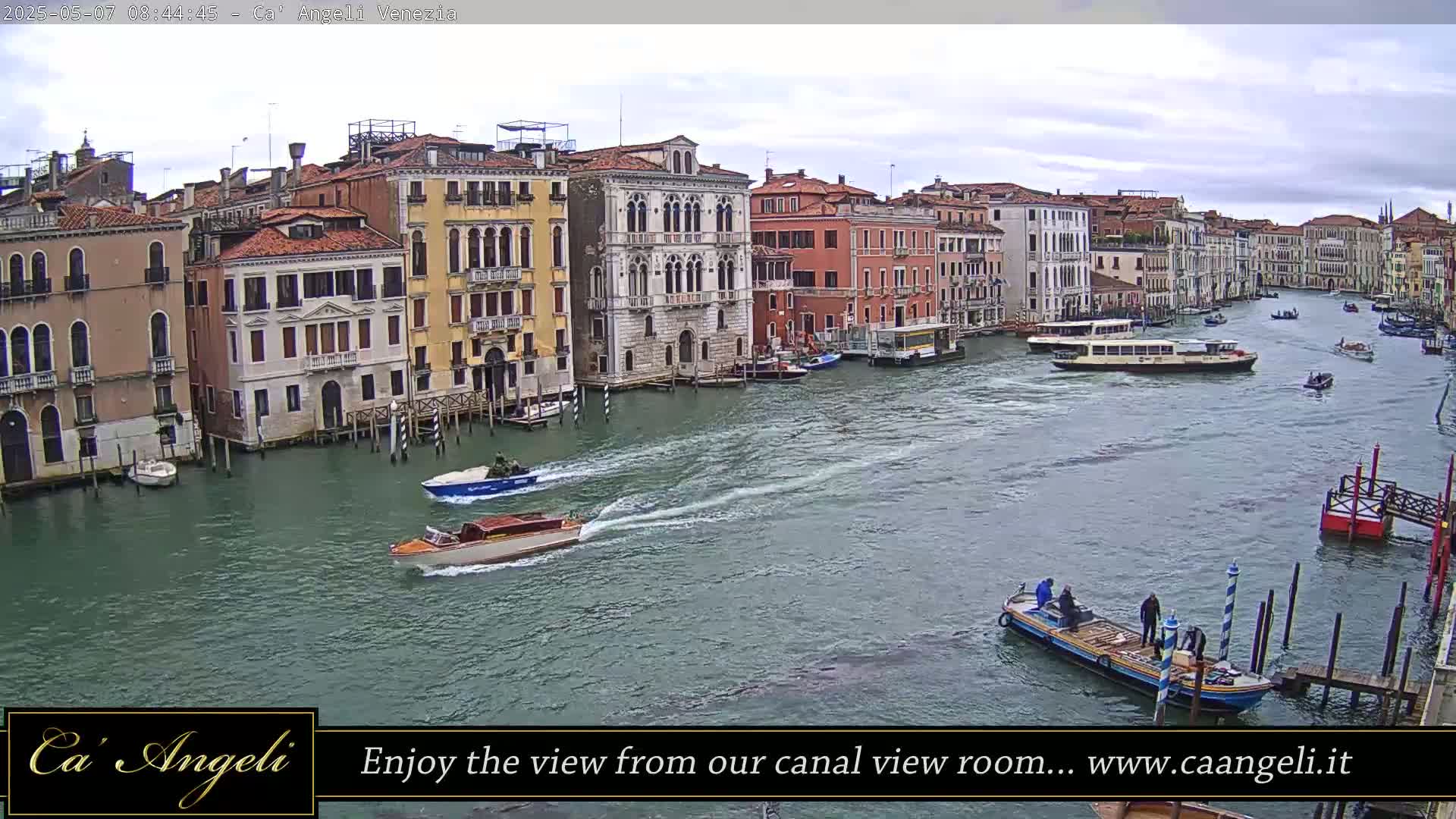A wide canal in Venice, under an overcast sky, is filled with various boats, with several colorful buildings lining its banks.