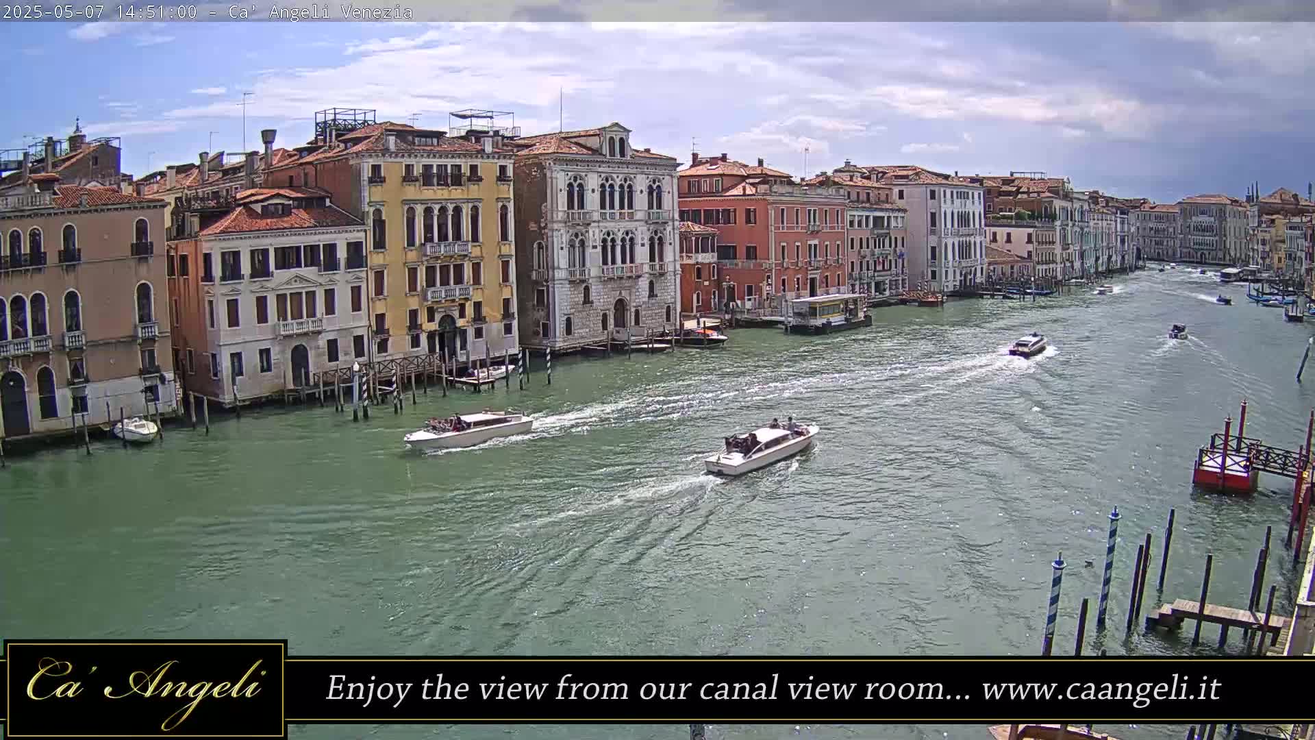 Several boats travel along a wide canal lined with closely spaced, multi-story buildings under a partly cloudy sky.