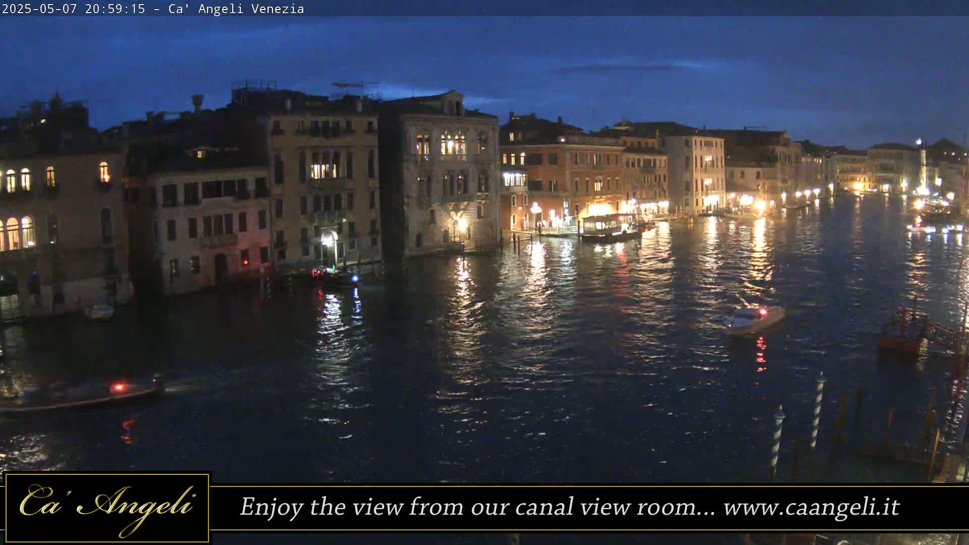 A nighttime view of a canal in Venice, Italy, with illuminated buildings lining the waterway and several boats on the water under a dark blue twilight sky.