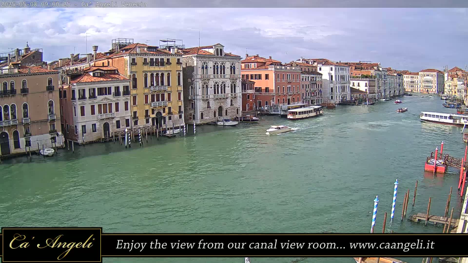 A wide shot of a canal in Venice, Italy, on a partly cloudy day, shows several buildings lining the waterway with boats traveling along it.