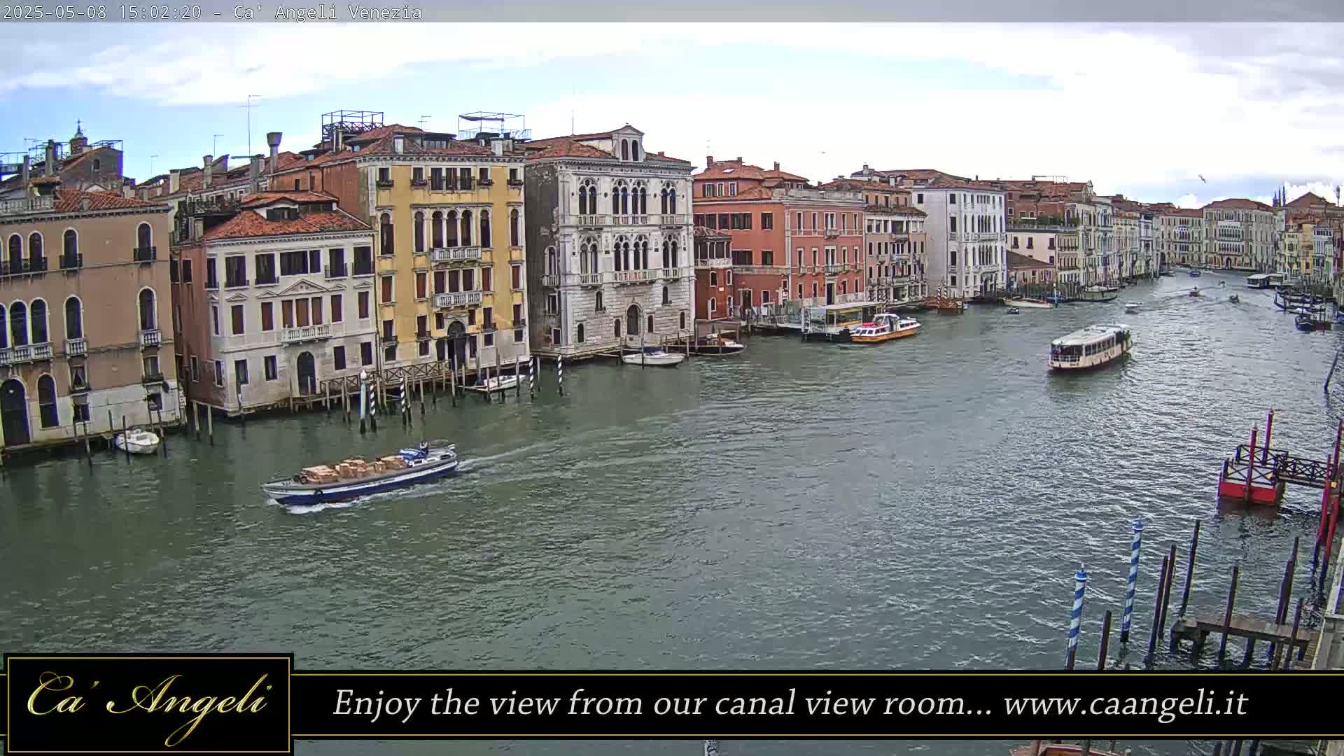 A wide shot of a Venetian canal on a partly cloudy day shows several boats and buildings lining the waterway.