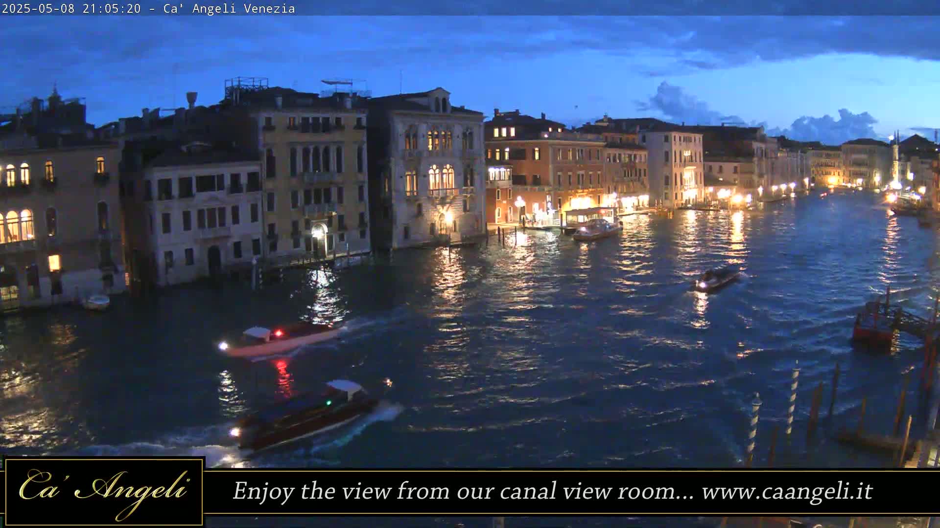 Under a twilight sky, several boats navigate a canal lined with illuminated buildings.