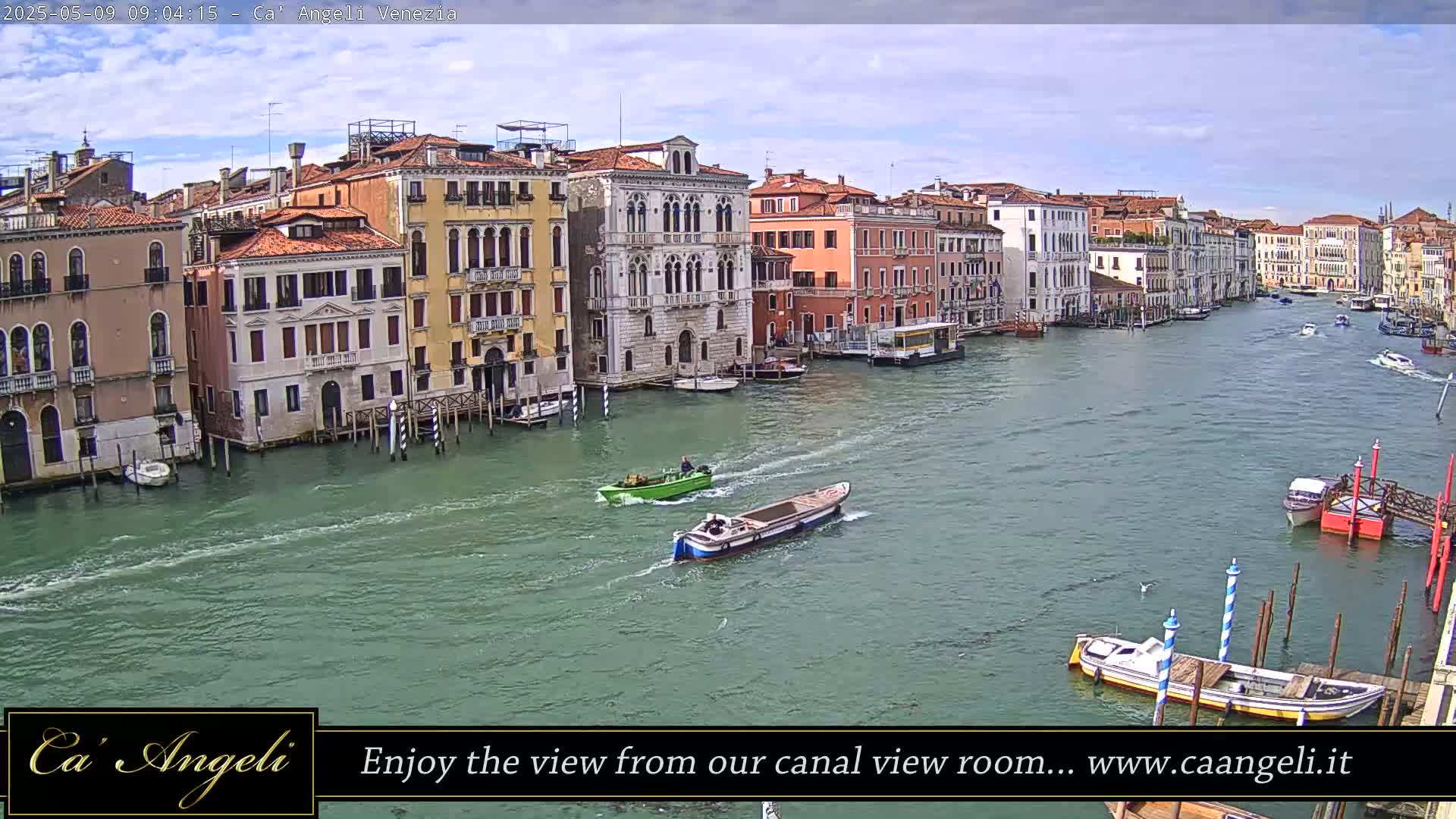 Under a partly cloudy sky, several boats traverse a wide canal lined with closely situated, multi-story buildings in Venice.