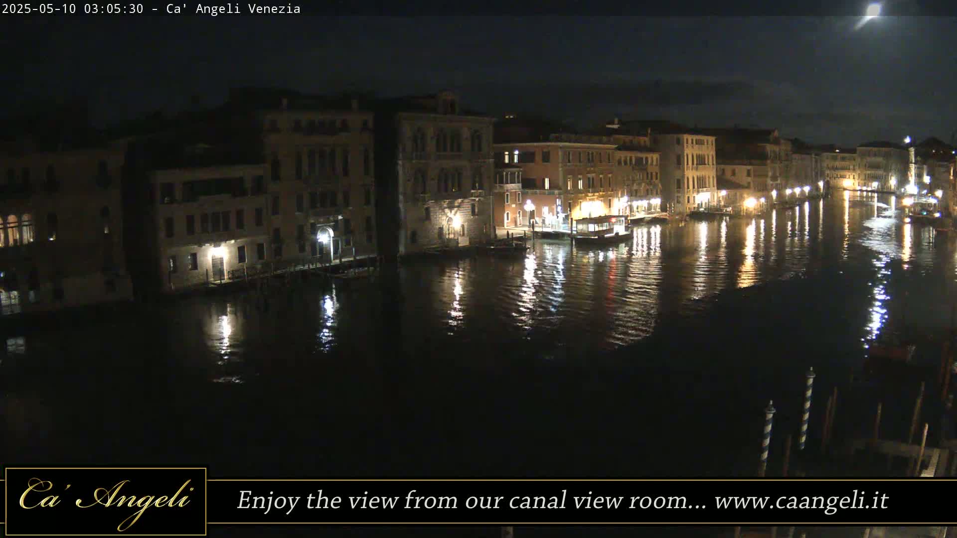 A nighttime view of a canal in Venice, Italy, shows illuminated buildings lining the waterway, their lights reflecting on the dark water.
