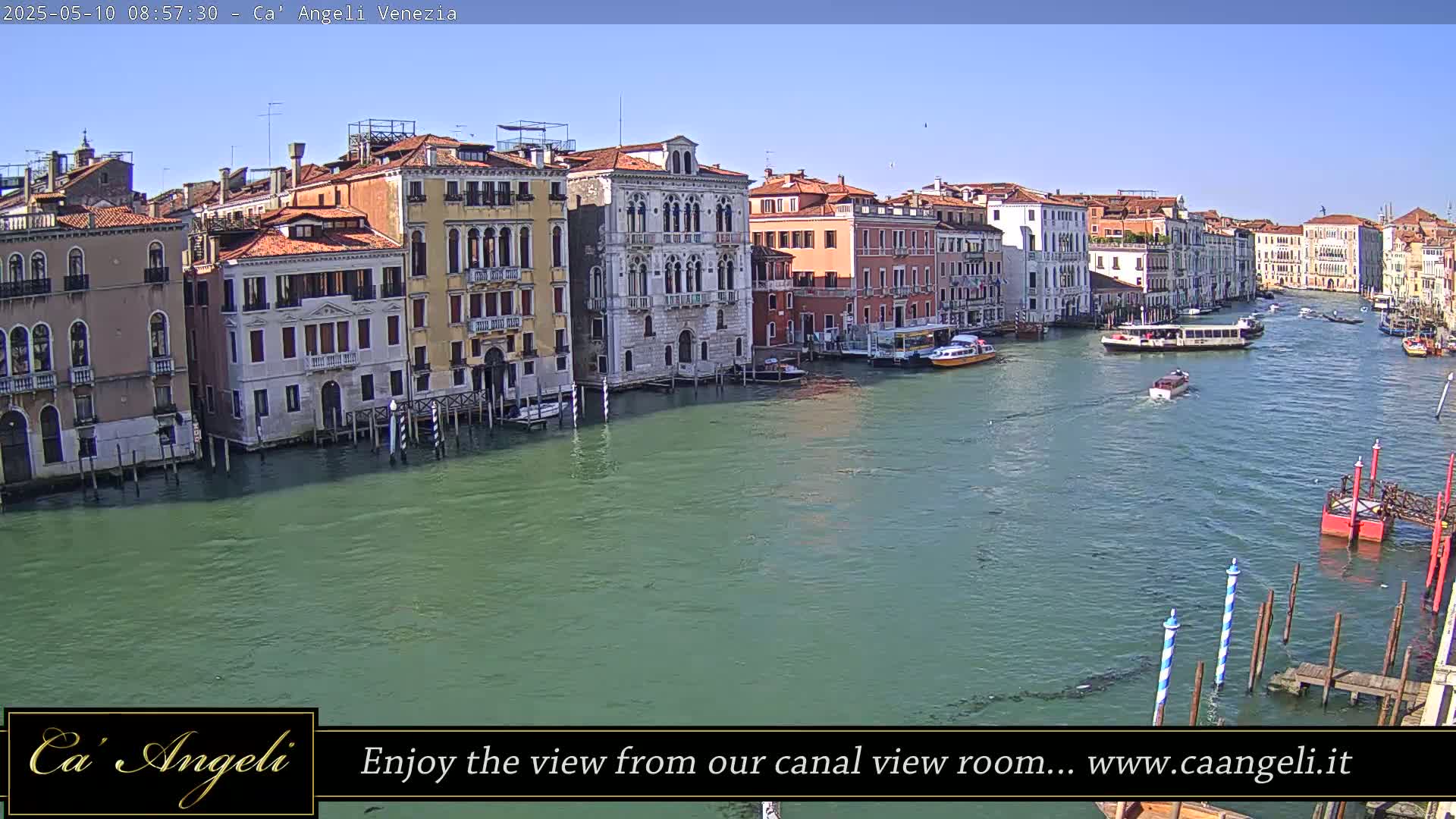 A sunny view of a Venetian canal with numerous buildings lining its banks, several boats on the water, and a small dock in the foreground.