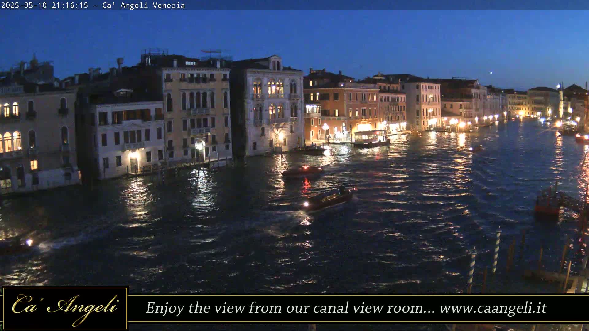 Several boats navigate a dark, calm canal at night, lined with illuminated buildings.