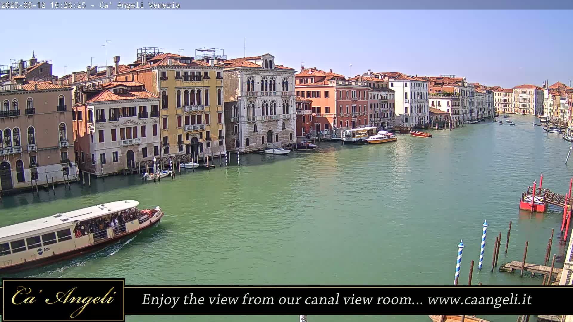 A waterway in Venice, Italy, shows numerous buildings lining the canal under a clear, sunny sky, with boats and a water bus traveling along the water.
