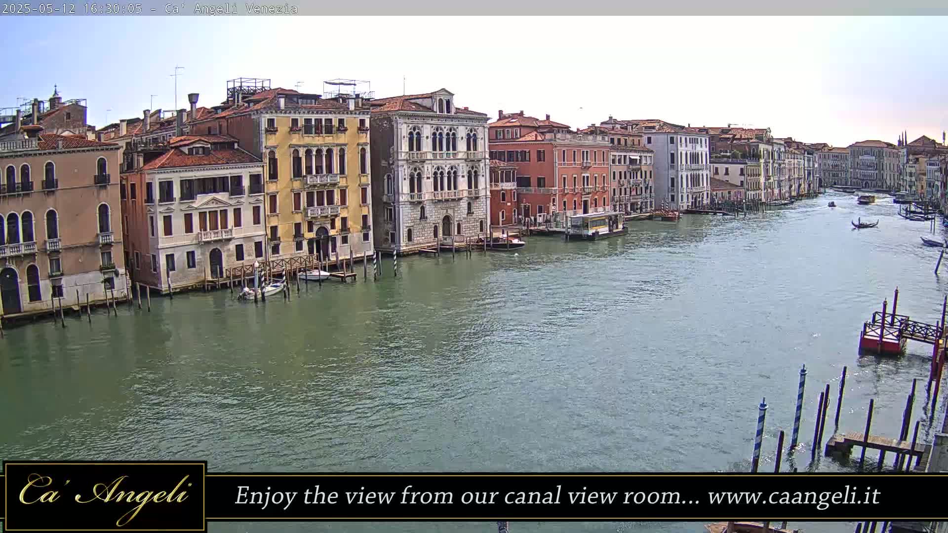 A wide shot of a canal in Venice, Italy, on a partly cloudy day, shows several buildings lining the waterway with boats and gondolas visible on the water.