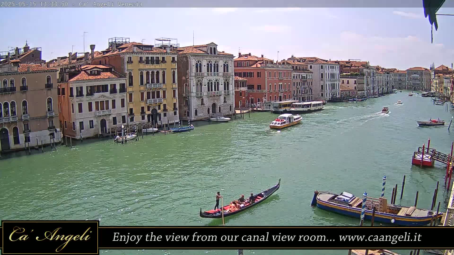 A gondola glides on a wide canal in Venice, lined with colorful buildings under a mostly clear sky.