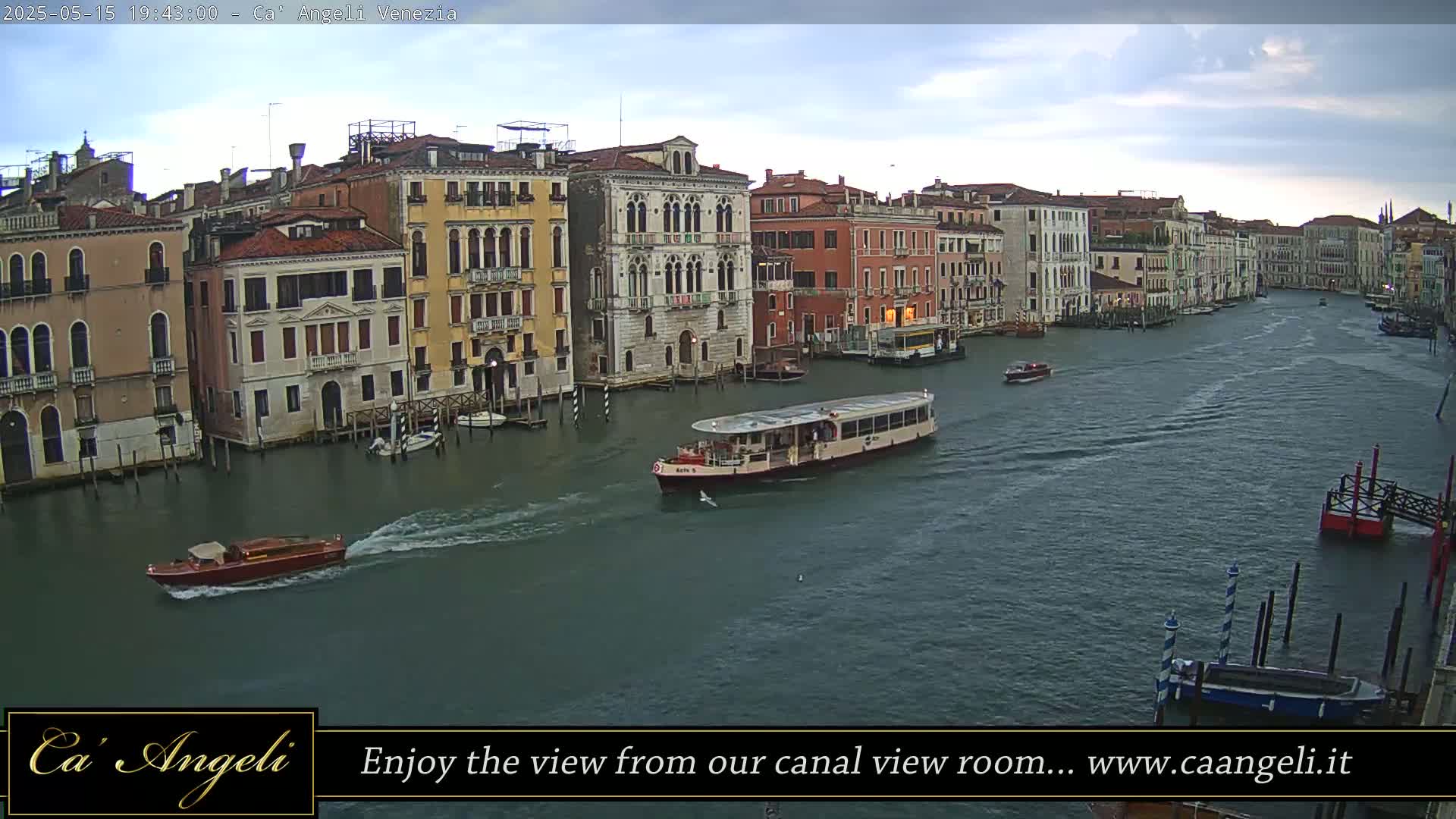 A wide canal in Venice, under a cloudy sky, is filled with several boats, including a large passenger ferry, with numerous buildings lining the waterway.
