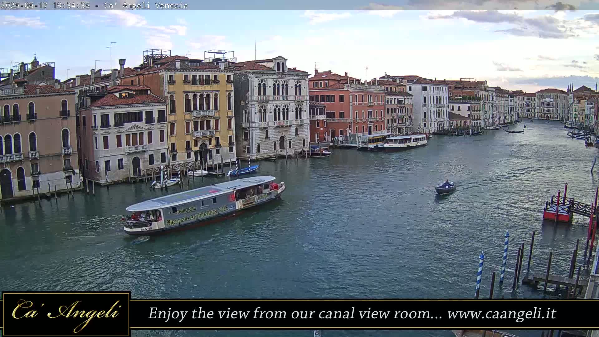 A wide canal in Venice, Italy, is shown under a partly cloudy sky, with several boats on the water and buildings lining the canal.