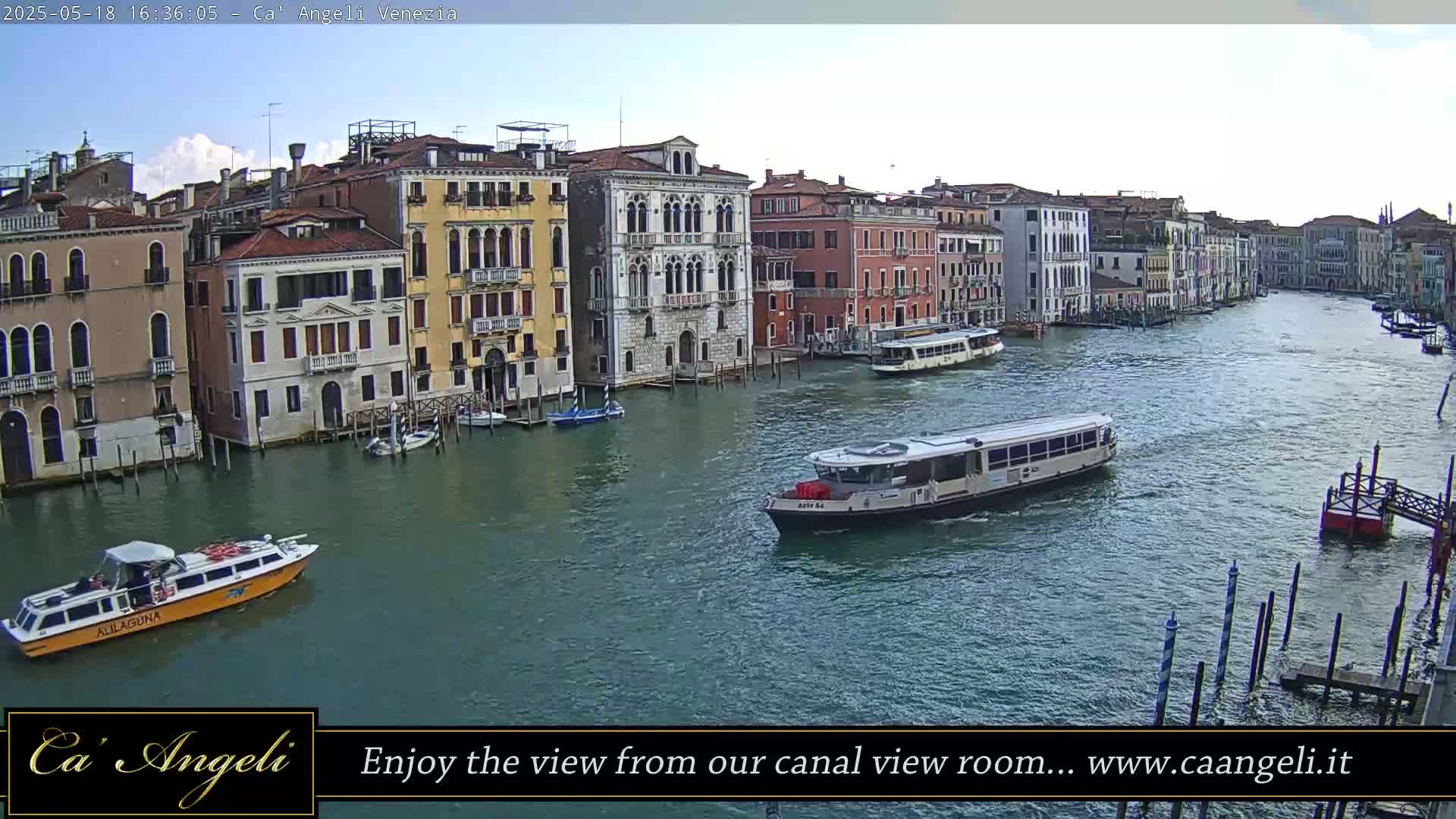 A waterway with several boats, including a water taxi, is lined with multi-story buildings under a mostly clear sky.