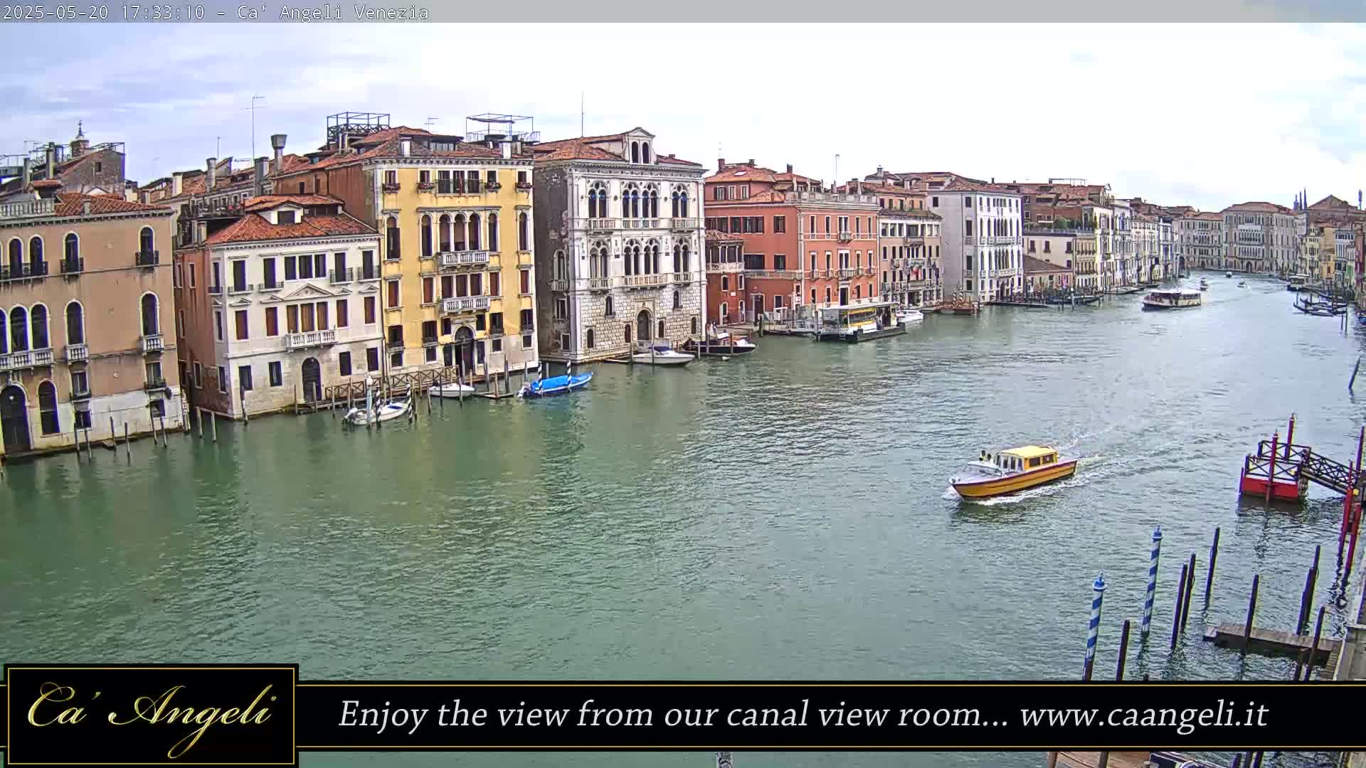 A wide shot of a Venetian canal on an overcast day shows colorful buildings lining the waterway, with several boats and water taxis traversing the calm water.