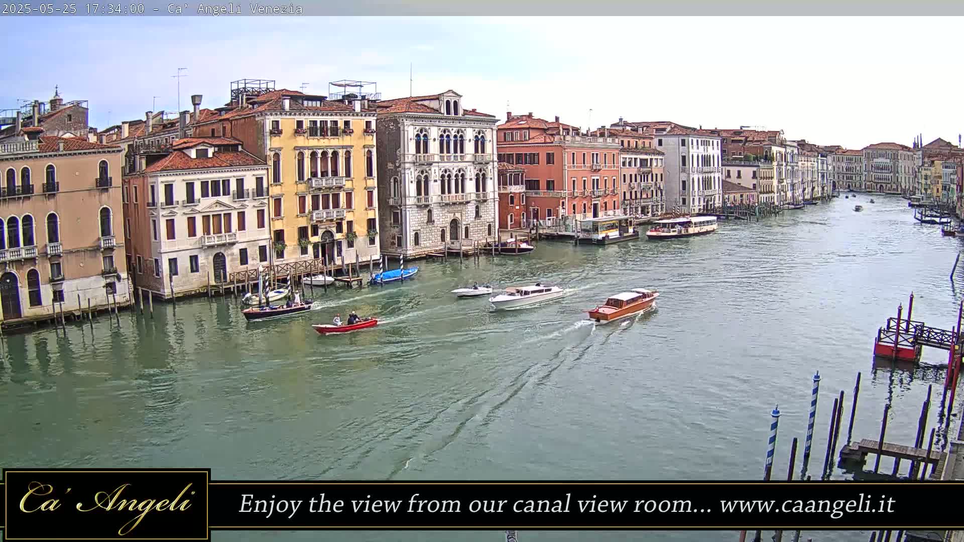 A wide shot of a canal in Venice, Italy, on a partly cloudy day, shows several boats and numerous buildings lining the waterway.