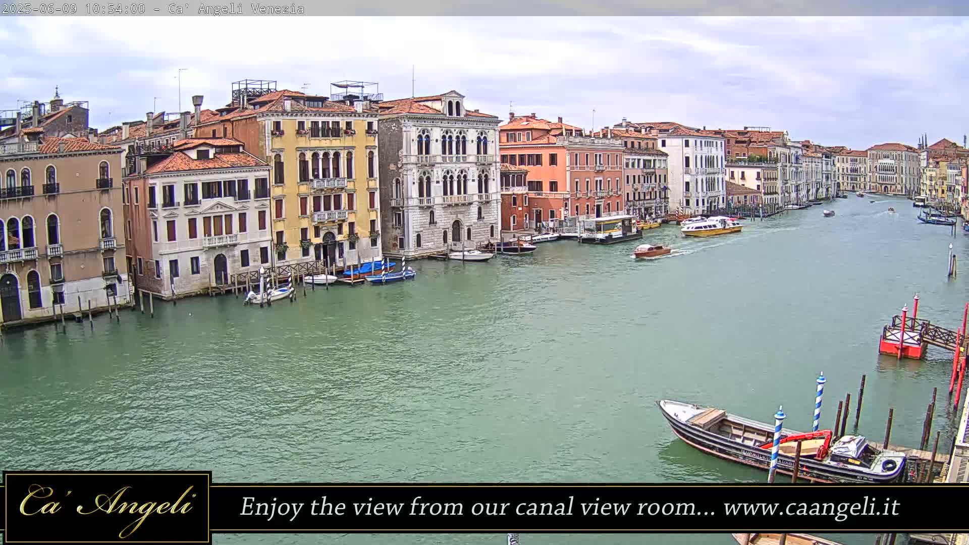 A wide shot of a Venetian canal on an overcast day shows numerous buildings lining the waterway, with several boats and a barge visible on the water.