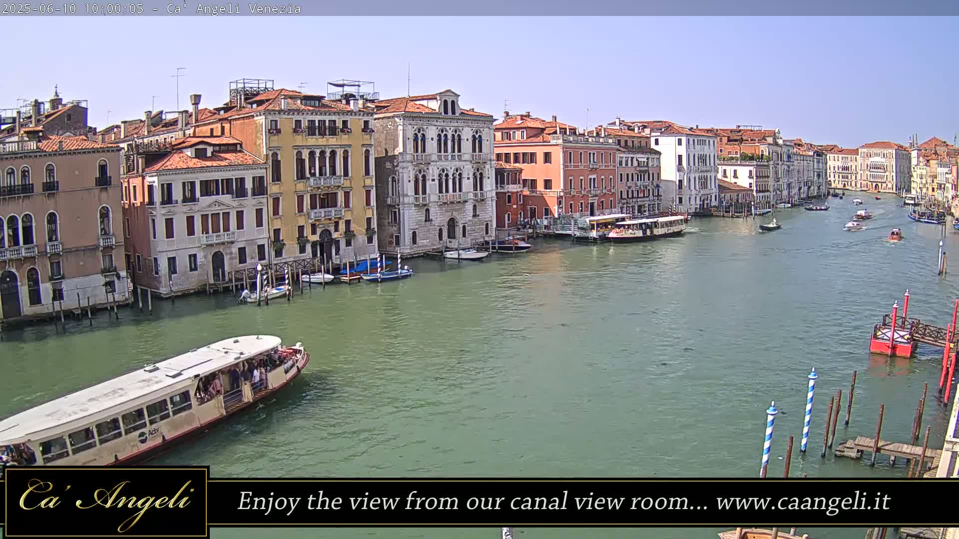 A wide shot of a Venetian canal on a sunny day shows several buildings lining the waterway, with boats including a water bus traveling along it.