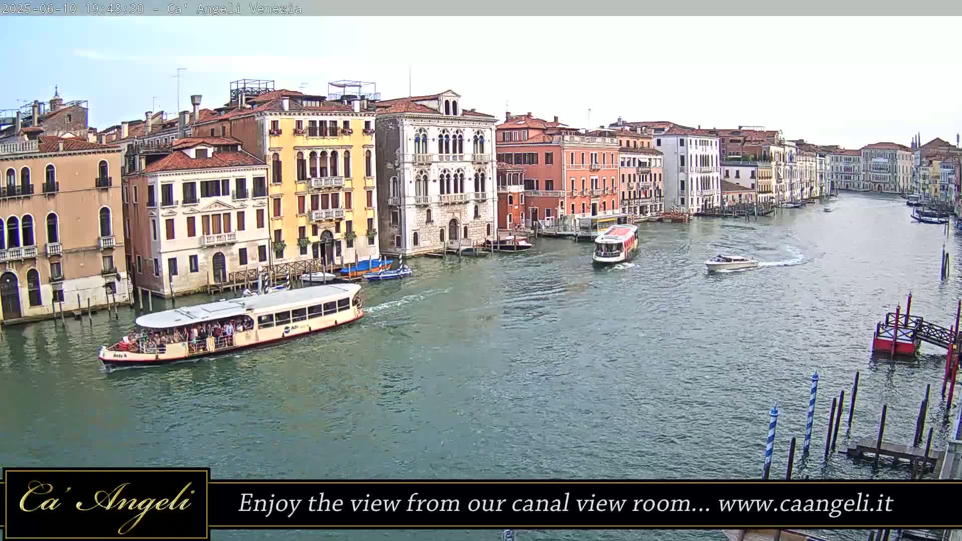 A wide canal in Venice, Italy is filled with several boats, under a mostly cloudy sky.