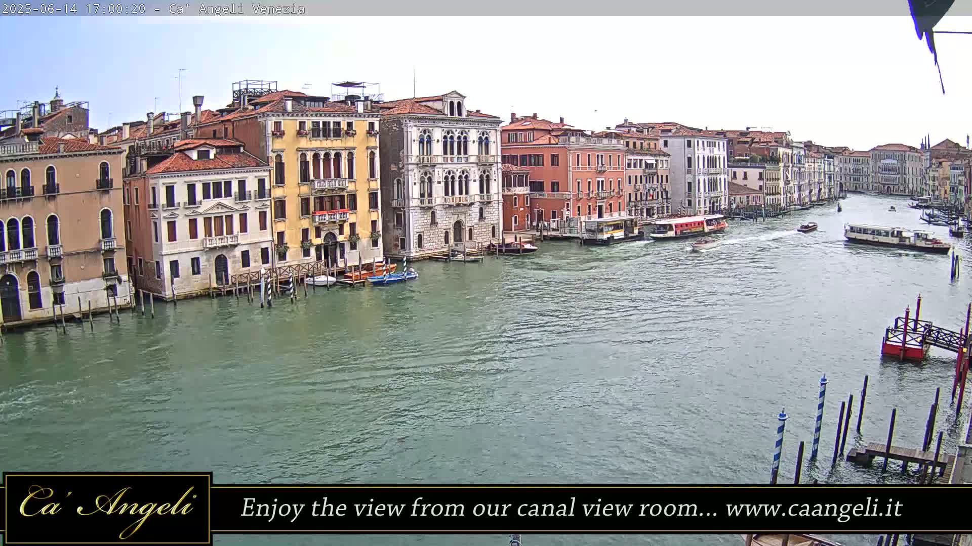 A wide shot of a waterway shows many buildings along the edge with boats passing by on a cloudy day.