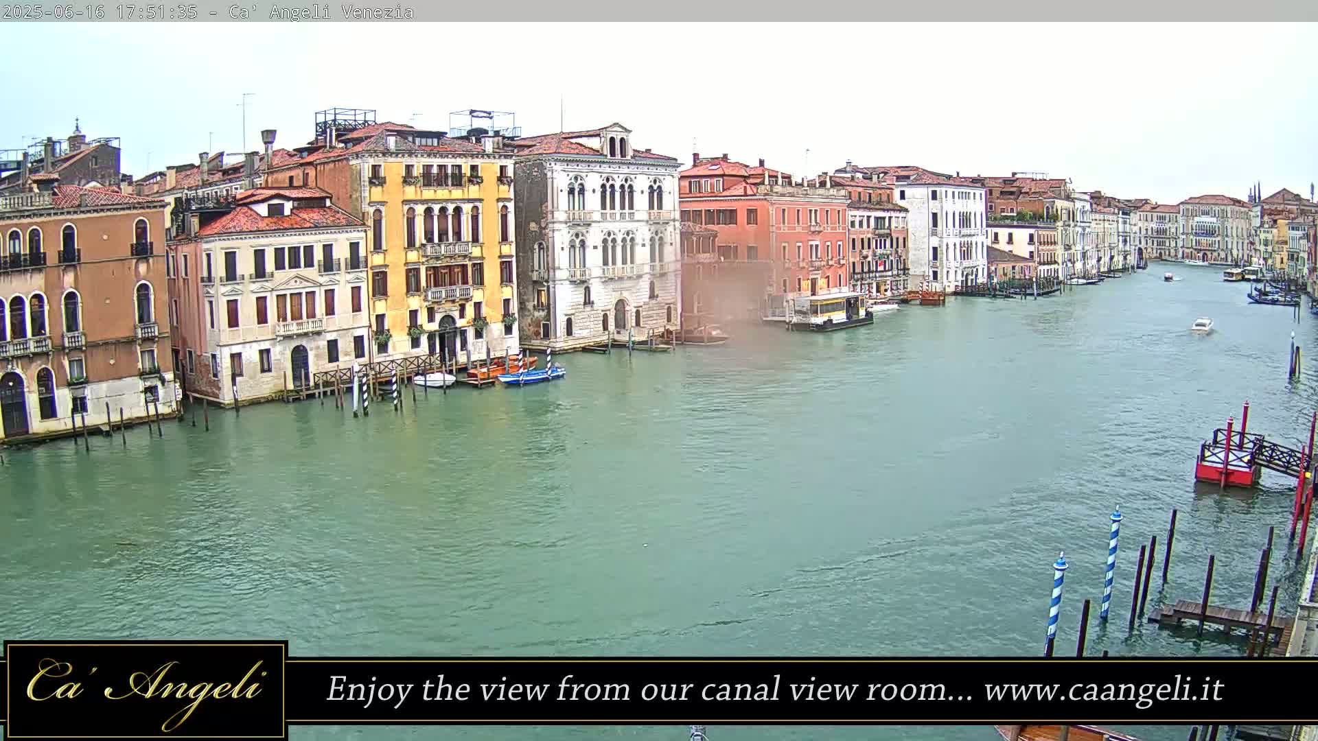 A wide shot of a Venetian canal shows several buildings lining the waterway under an overcast sky, with a few boats and mooring poles visible in the water.